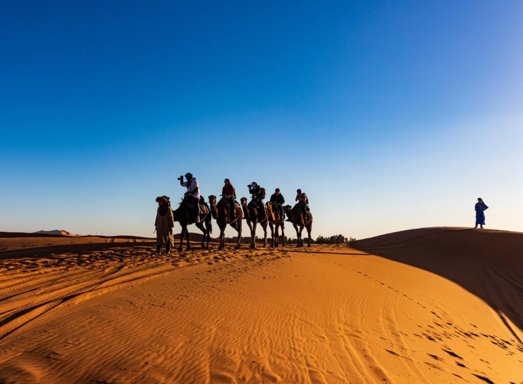 A group of travelers on camels crossing the sandy dunes of the Sahara Desert under a clear blue sky.