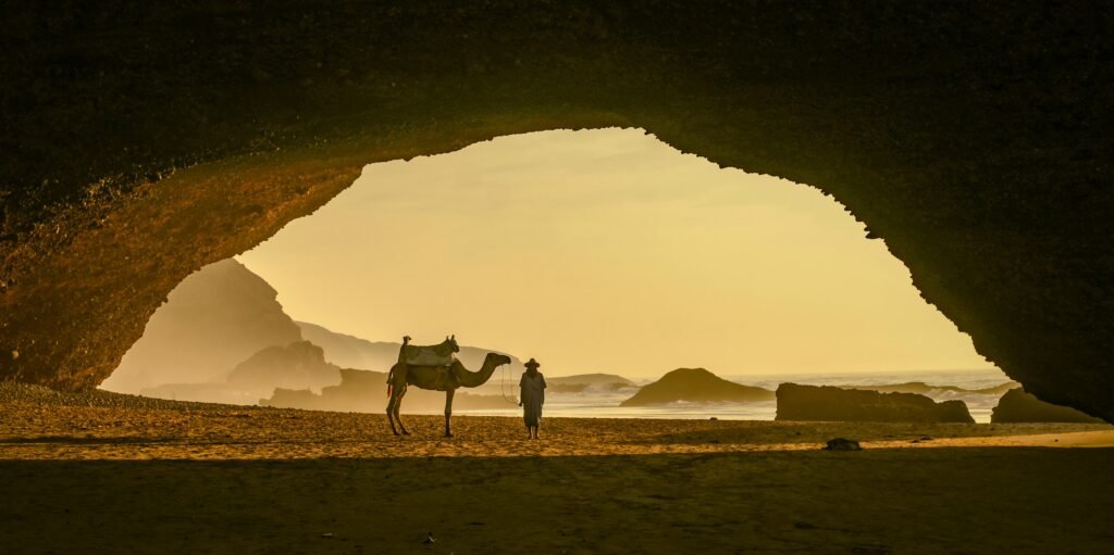 Silhouetted scene of a camel and person at sunset under the iconic Legzira Beach rock arch.